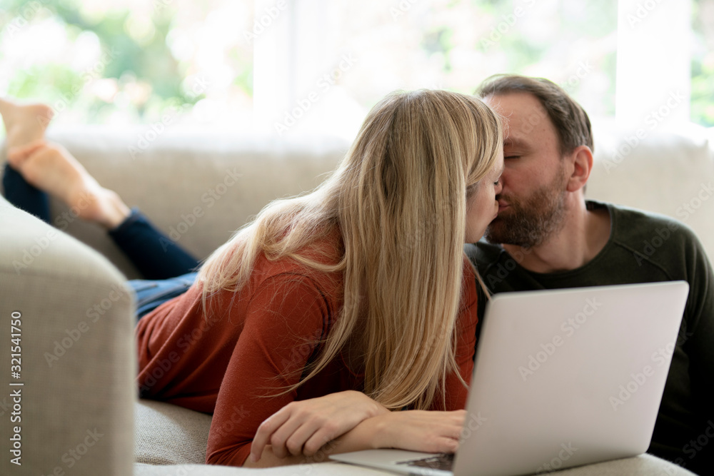 Couple sitting at home on couch, kissing, using laptop Couple sitting at home on couch, kissing, using laptop
