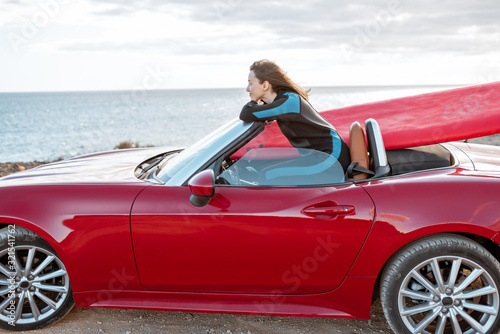 Woman enjoying beautiful landscapes while sitting on her sports car with a surfboard near the ocean. Carefree lifestyle and active sports concept