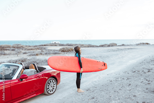 Portrait of a young woman surfer in swimsuit standing with surfboard on the dirt road near the ocean. Active lifestyle and surfing concept photo