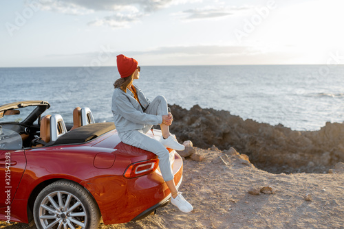 Woman enjoying beautiful view on the ocean, sitting on the convertible car on the rocky coast during a sunset. Carefree lifestyle and car travel concept