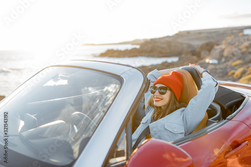 Lifestyle portrait of happy woman in red hat driving convertible car on the rocky ocean coast during a sunset. Carefree lifestyle and car travel concept