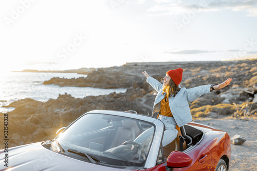 Woman enjoying beautiful view on the ocean, sitting with raised hands on the convertible car during a sunset. Wide view with space on the sky