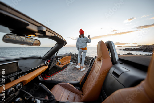 Woman enjoying beautiful view on the ocean, standing on the rocky coast during a sunset, view from the vehicle interior. Nature enjoyment and car travel concept