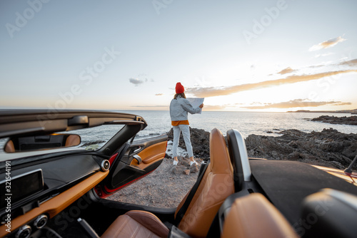 Woman enjoying beautiful view on the ocean, standing with map on the rocky coast during a sunset, wide view from the vehicle interior. Nature enjoyment and car travel concept