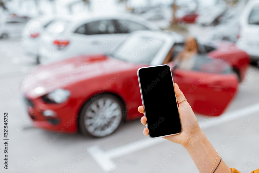 Woman holding a smartphone with empty screen at the car parking with red sports vehicle on the background, close-up view
