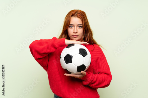 Teenager redhead girl with sweater over isolated green background holding a soccer ball