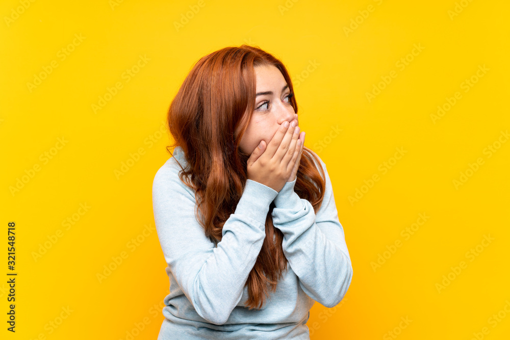 Teenager redhead girl over isolated yellow background covering mouth and looking to the side