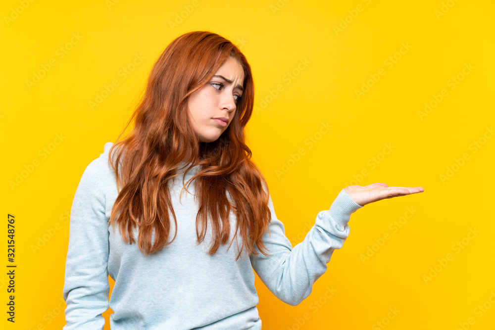 Teenager redhead girl over isolated yellow background holding copyspace with doubts Teenager redhead girl over isolated yellow background holding copyspace with doubts