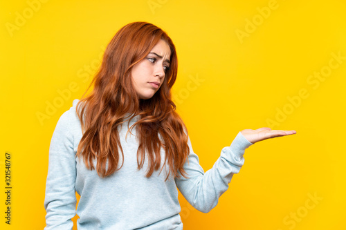 Teenager redhead girl over isolated yellow background holding copyspace with doubts