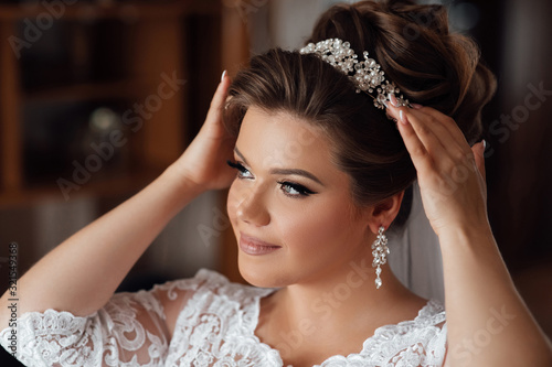 Beautiful bride in white dress with hairstyle and makeup. Bride touching crown. Stylis bride, wedding morning preparation. Bridal getting ready. Charming bride in white wedding dress.