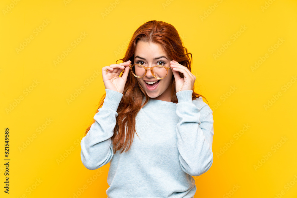 Teenager redhead girl over isolated yellow background with glasses and surprised - obrazy, fototapety, plakaty 