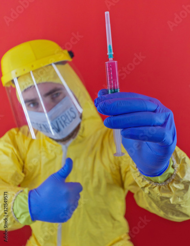 Man in yellow chemical protection suit and face protective mask  with the inscription `coronavirus`. Shoulder portrait. Red background. Without glasses. Protective helmet. Hands in blue gloves. 