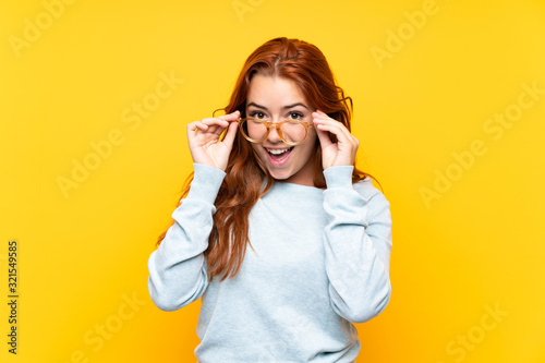 Teenager redhead girl over isolated yellow background with glasses and surprised photo