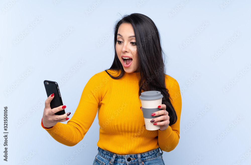 Young Colombian girl over isolated blue background holding coffee to take away and a mobile