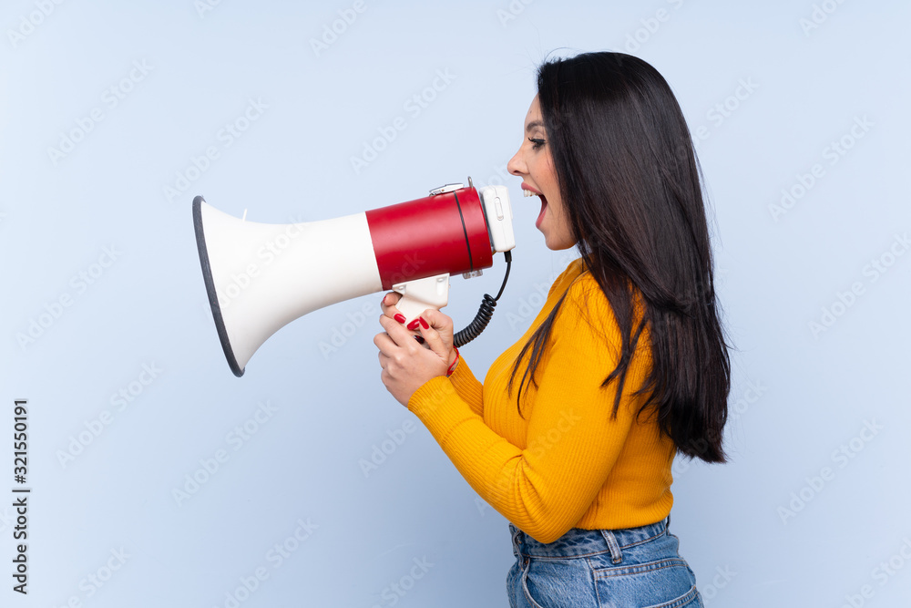 Young Colombian girl over isolated blue background shouting through a megaphone Young Colombian girl over isolated blue background shouting through a megaphone