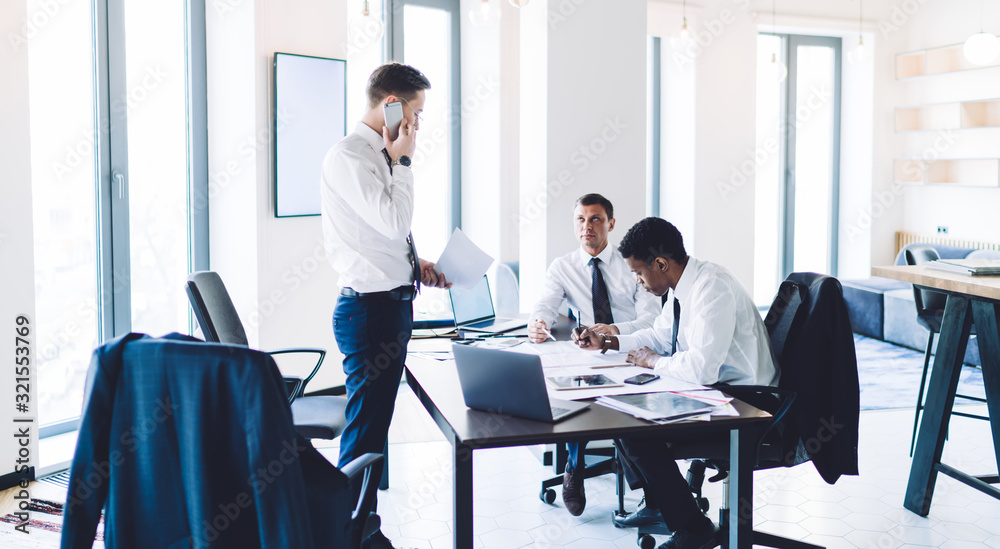 Businessman with papers standing and phoning while working with multiethnic colleagues in office Businessman with papers standing and phoning while working with multiethnic colleagues in office