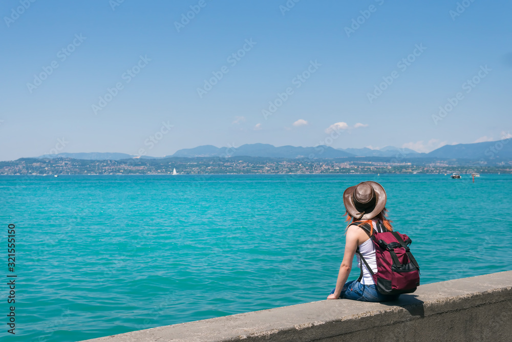 A girl in a cowboy hat and a backpack sits alone on the promenade, looks at the turquoise water of Lake Garda. Vacation. Travels and tours to Europe. Sunny summer day. Italy. copy space