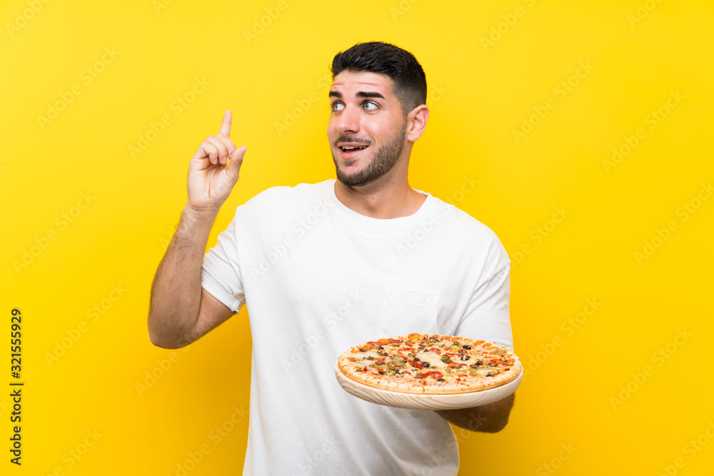 Young handsome man holding a pizza over isolated yellow wall intending to realizes the solution while lifting a finger up Young handsome man holding a pizza over isolated yellow wall intending to realizes the solution while lifting a finger up