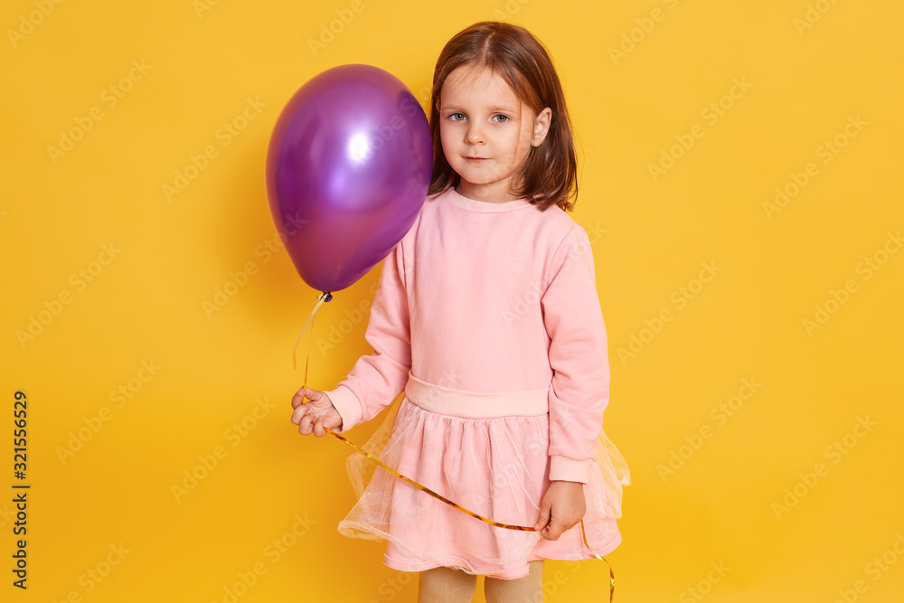 Horizontal indoor picture of sweet positive good looking little girl standing isolated over yellow background, holding air baloon in one hand, looking directly at camera, wearing pink dress. Horizontal indoor picture of sweet positive good looking little girl standing isolated over yellow background, holding air baloon in one hand, looking directly at camera, wearing pink dress.