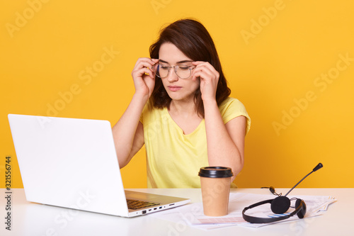 Studio shot of sitting near white lap top and take away coffe, touching her eyewear, looks tired, working online, charming brunette lady posing isolated over yellow background. Technology cocnept. photo