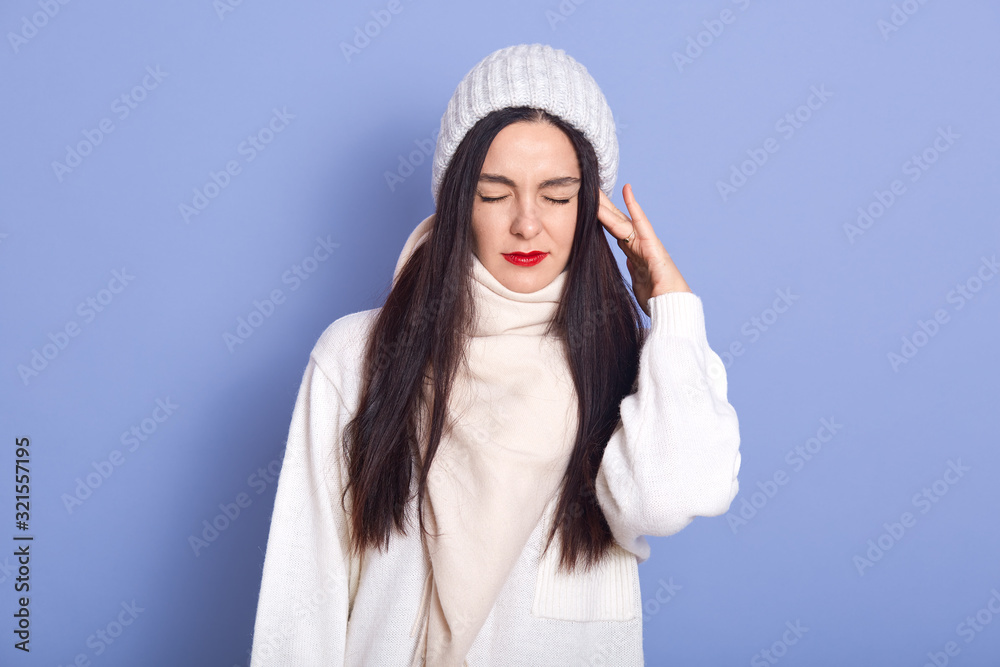 Horizontal picture of young attractive woman standing isolated over blue background in studio, putting hand on ear, having earache, having caught cold, closing eyes, wearing white warm clothes. Horizontal picture of young attractive woman standing isolated over blue background in studio, putting hand on ear, having earache, having caught cold, closing eyes, wearing white warm clothes.