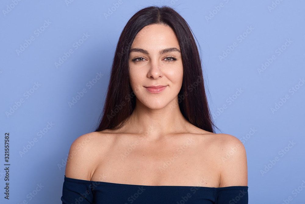 Naklejka premium Studio shot of pretty pleased woman smiles positively at camera, being in high spirit, having healthy skin, wearing dress or shirt, demonstrates bare shoulders, models against blue background.