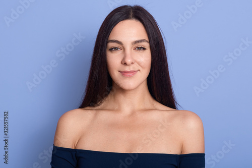 Studio shot of pretty pleased woman smiles positively at camera, being in high spirit, having healthy skin, wearing dress or shirt, demonstrates bare shoulders, models against blue background. photo