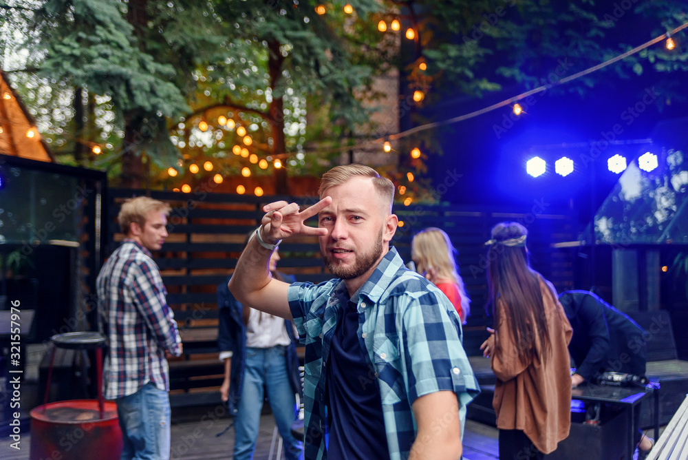 Handsome 30s caucasian young man dancing on near the camera on the background of his friends at the party in cozy evening garden.