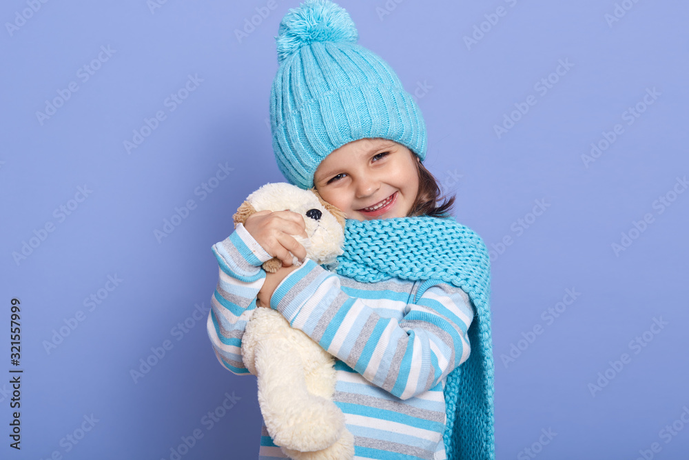 Horizontal photo of cute little girl dressed cap, scarf and stripes shirt, holding plush toys up to her face, looking directly at camera withcharming smile, having positive emotions. Chidhood concept. Horizontal photo of cute little girl dressed cap, scarf and stripes shirt, holding plush toys up to her face, looking directly at camera withcharming smile, having positive emotions. Chidhood concept.