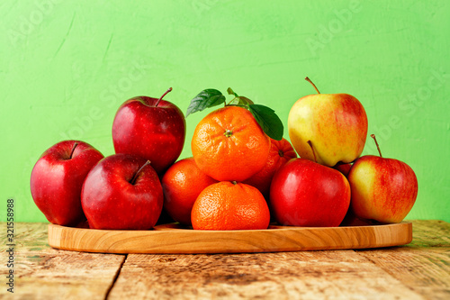 Red ripe apples and tangerines with green leaves lie on a wooden tray on an old wooden table with light green background.