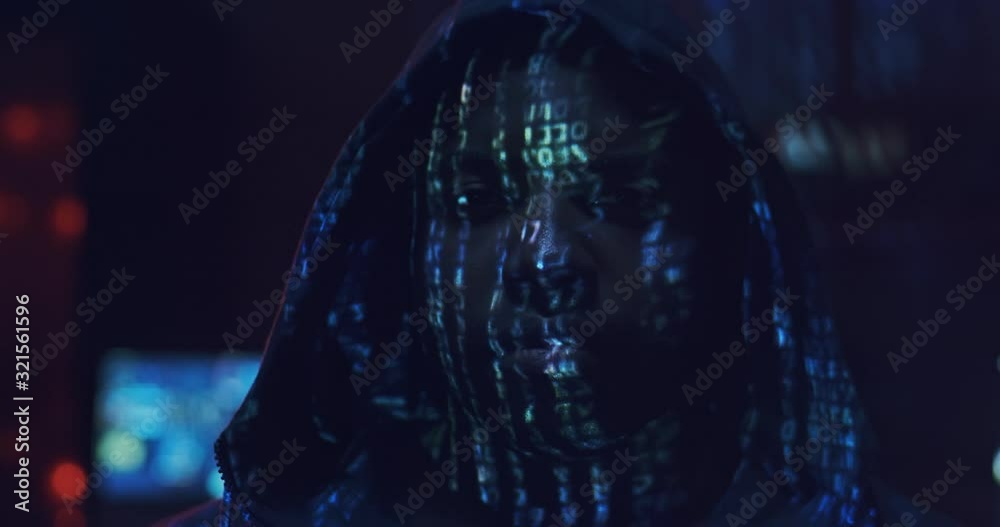 Close up of the young African American guy hacker in a hood looking straight to the camera with projected code numbers and characters on His Face in the dark room full of computers. Portrait. Close up of the young African American guy hacker in a hood looking straight to the camera with projected code numbers and characters on His Face in the dark room full of computers. Portrait.