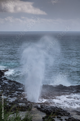 Koloa, Kauai, Hawaii, USA. - January 16, 2020: Gray-azure Pacific Ocean with black rocky coastline shows eruption of the Spouting Horn geiser under cloudscape.