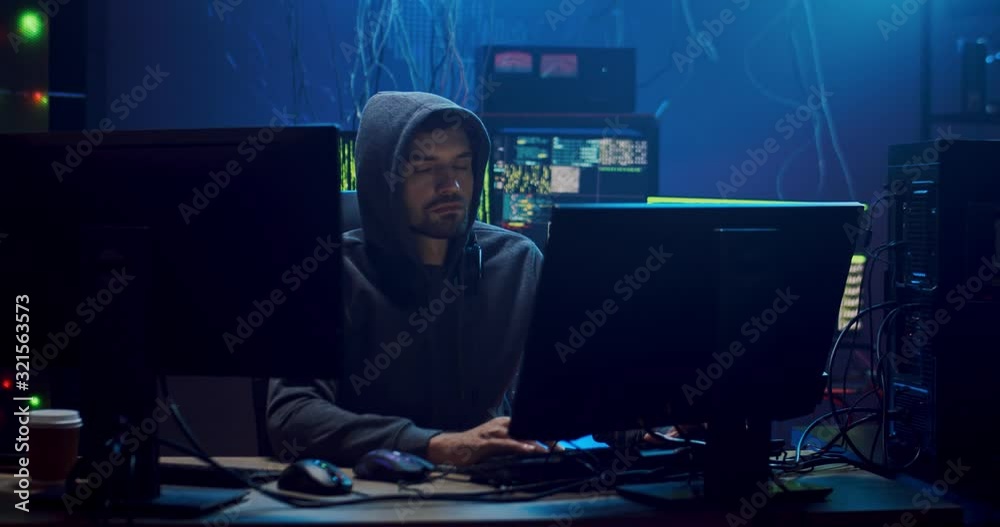 Male Caucasian hacker in a hood programming at the two big screen of ...