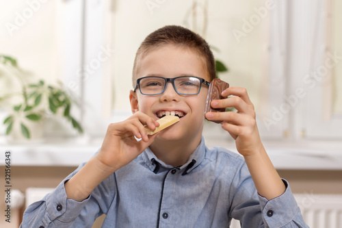 Uśmiechnięty chłopiec w okularach siedzi przy stole i trzyma w dłoniach słodycze. W jednej ręce trzyma białą czekoladę którą gryzie a w drugiej czekoladowego batona.