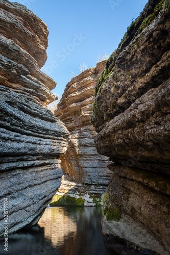 Beautiful view through the narrow, rocky, slot canyon, reflective mountain creek water and vertical cliffs covered by green moss under a blue sky