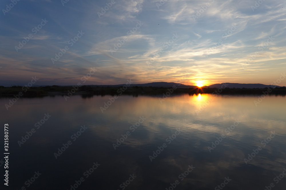 Sunset reflection in lake with mountains in the horizon, calm water and small stringy clouds in blue sky. Yellow sun on edge of mountain with pink and blue colors