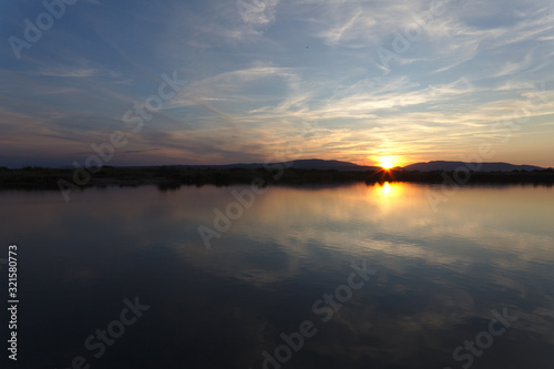 Sunset reflection in lake with mountains in the horizon  calm water and small stringy clouds in blue sky. Yellow sun on edge of mountain with pink and blue colors