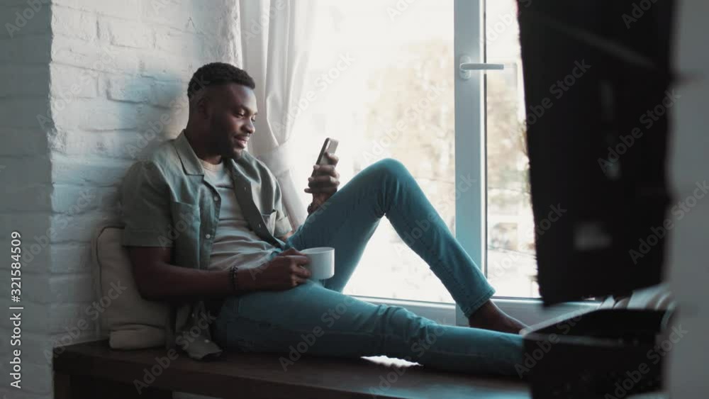 Handsome african american young man in the modern apartment sitting on windowsill looking through the window drinking cofee holding and using a smartphone Handsome african american young man in the modern apartment sitting on windowsill looking through the window drinking cofee holding and using a smartphone