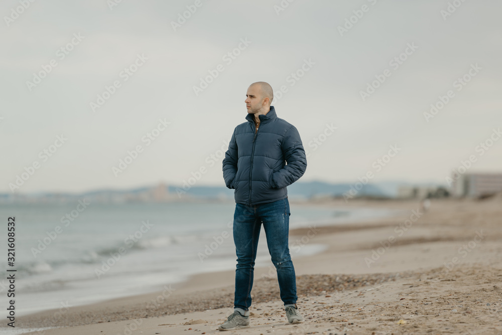 A muscular bald man in the jeans, bologna jacket and sneakers poses near the Mediterranean Sea