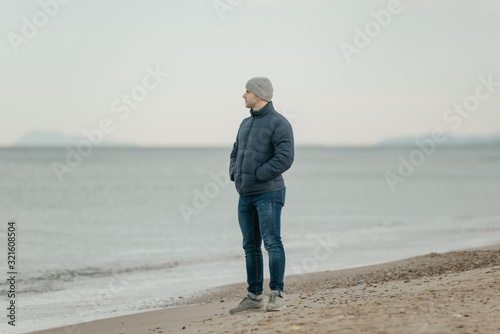 A muscular man in the grey hat, jeans, bologna jacket, and sneakers looks in the Mediterranean Sea