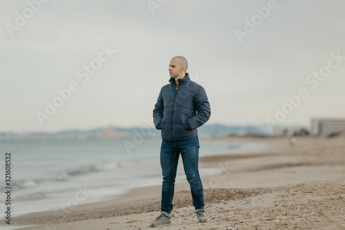 A muscular bald man in the jeans, bologna jacket and sneakers poses near the Mediterranean Sea