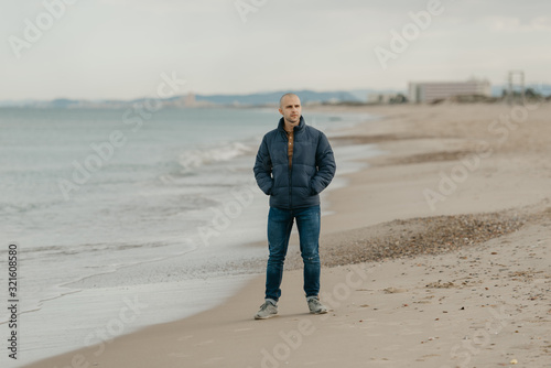 A muscular bald stylish man in the jeans, bologna jacket and sneakers waits with hands in the pockets near the Mediterranean Sea