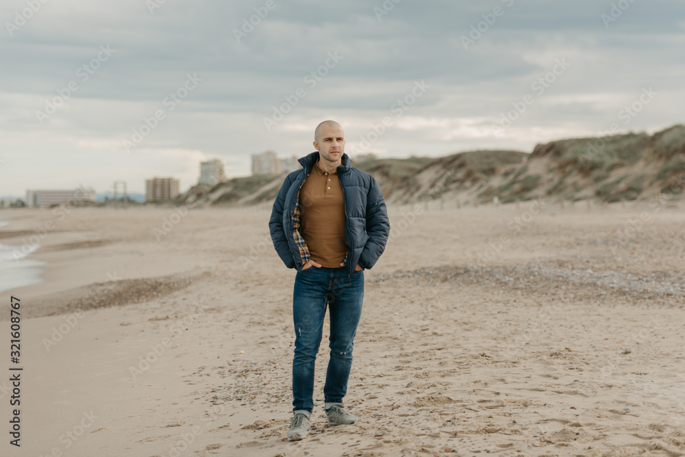 A muscular bald stylish man in the down jacket, jeans and sneakers walks with hands in jeans pockets near the Mediterranean Sea in the afternoon