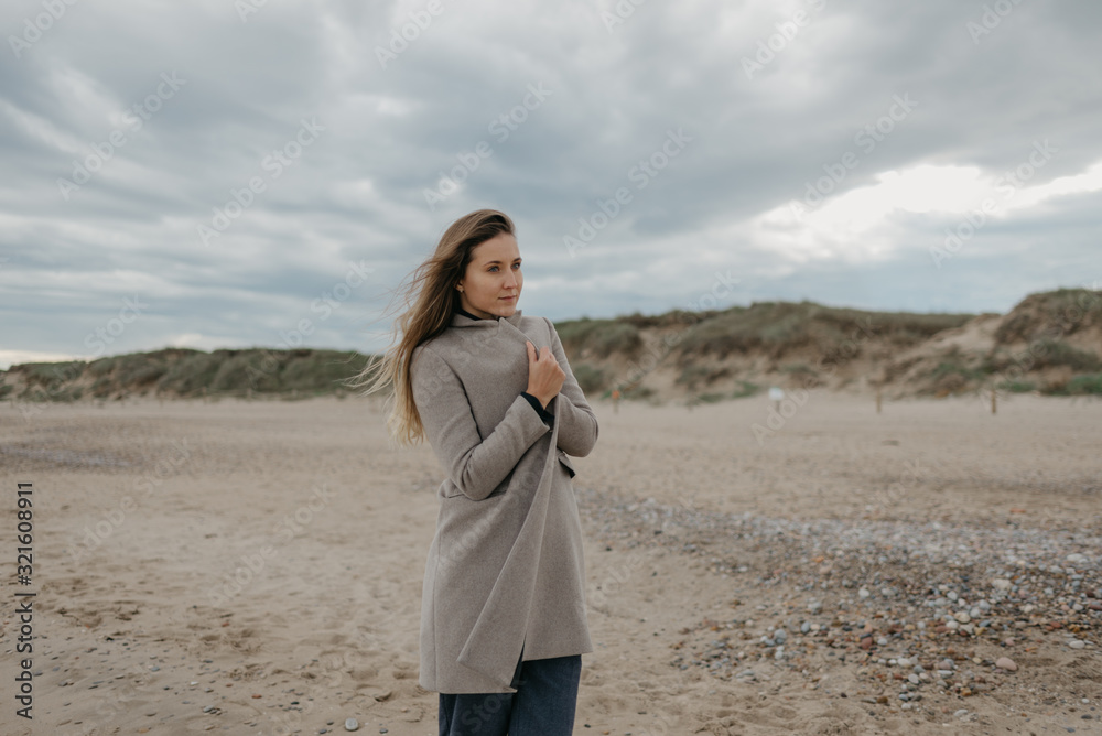 A gorgeous attractive woman with long hair with a smile wraps herself in a coat near The Mediterranean Sea. Sandy dunes in the background. A gorgeous attractive woman with long hair with a smile wraps herself in a coat near The Mediterranean Sea. Sandy dunes in the background.