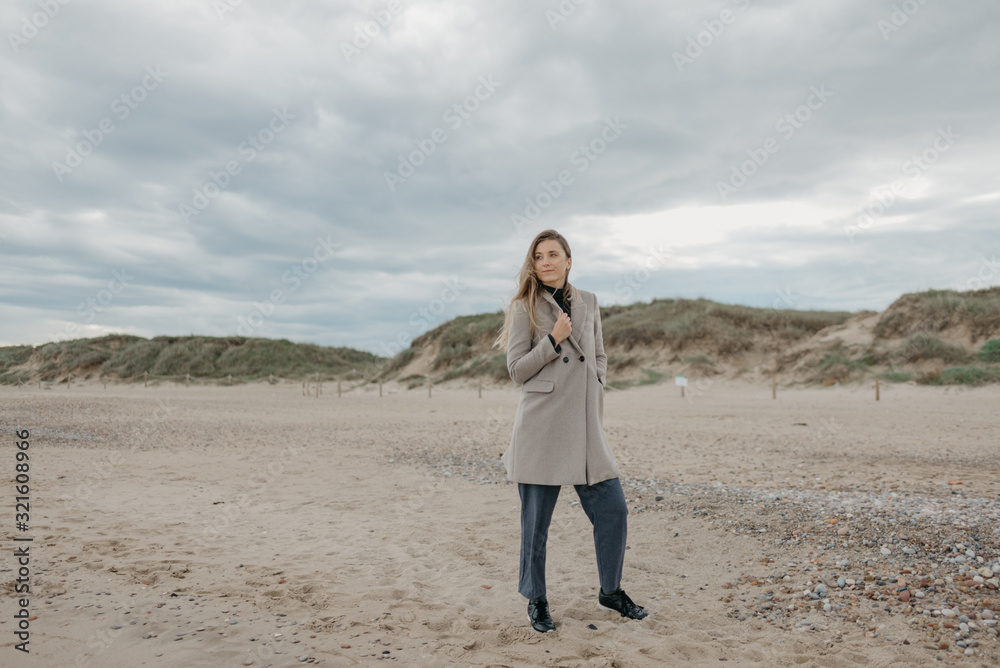 A gorgeous attractive woman with long hair wraps herself in a coat on a shore of the Mediterranean Sea. Sandy dunes in the background. A gorgeous attractive woman with long hair wraps herself in a coat on a shore of the Mediterranean Sea. Sandy dunes in the background.