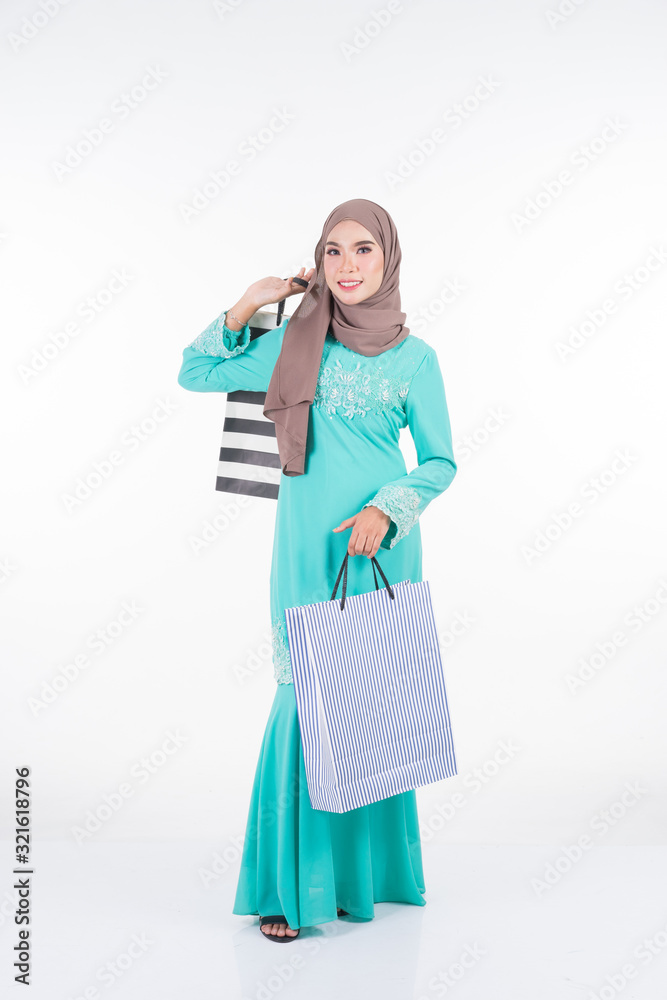 A beautiful and excited Muslim female model in a Asian traditional dress modern kurung carrying shopping bags isolated on white background. Eidul fitri festive shopping concept. Full length portrait.