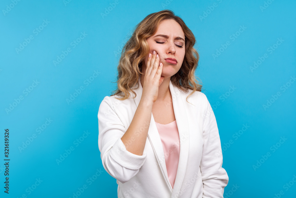 Toothache. Portrait of unhappy sick woman with wavy hair in white jacket suffering dental pain, standing with closed eyes and touching cheek to relieve pain. studio shot isolated on blue background Toothache. Portrait of unhappy sick woman with wavy hair in white jacket suffering dental pain, standing with closed eyes and touching cheek to relieve pain. studio shot isolated on blue background