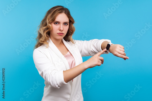 Times up! Portrait of serious woman with wavy hair in white jacket pointing at watch on hand and looking disapprovingly, dissatisfied with being late. indoor studio shot isolated on blue background photo