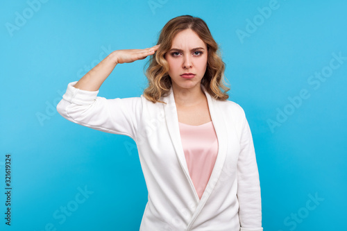Yes sir! Portrait of serious woman with wavy hair in white jacket giving salute, obediently listening to commander order with attentive confident face. indoor studio shot isolated on blue background