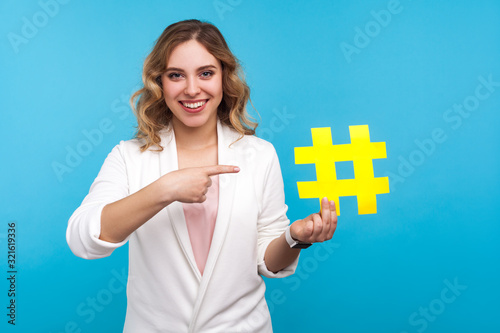Hashtag and blogging. Portrait of beautiful cheerful woman with wavy hair in white jacket pointing at big paper hash symbol and smiling, internet popularity. studio shot isolated on blue background